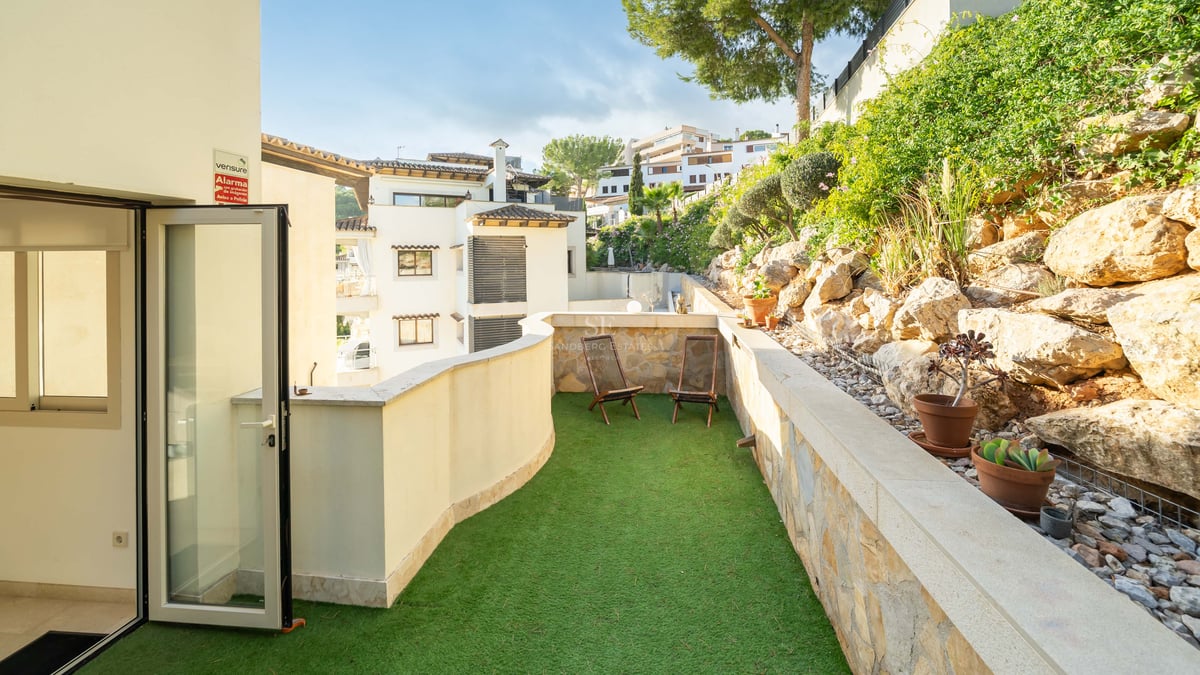 Terrasse ensoleillée avec gazon artificiel vert, deux chaises en bois et un mur en pierre naturelle.