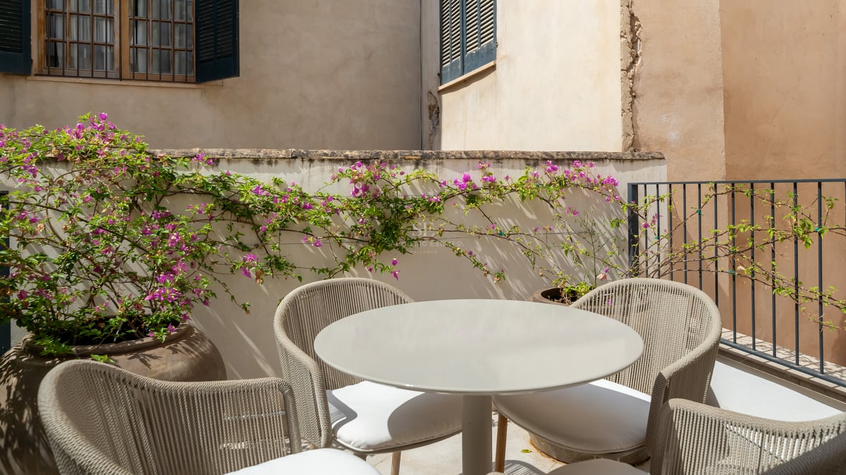 Mesa redonda gris y sillas de cuerda trenzada en una terraza soleada con buganvillas rosas y muros de piedra.