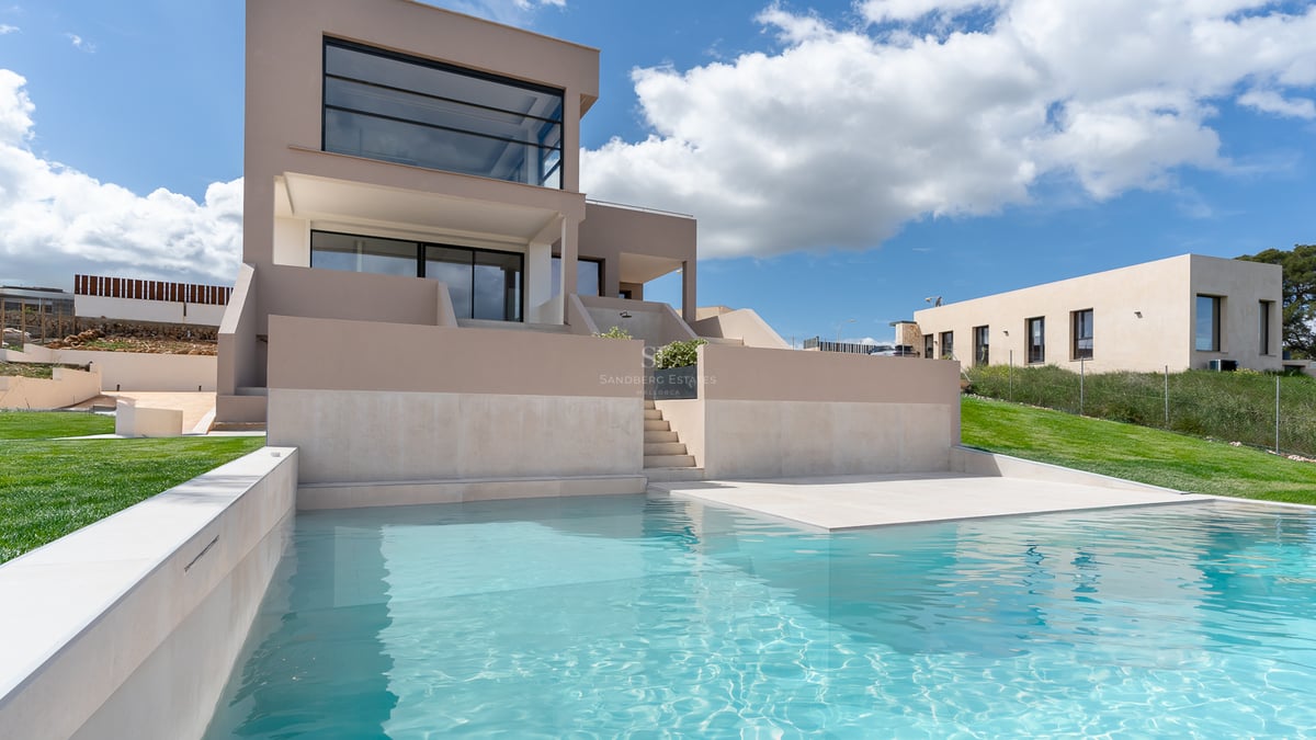 Piscine turquoise au premier plan devant une villa contemporaine beige sous un ciel bleu parsemé de nuages.