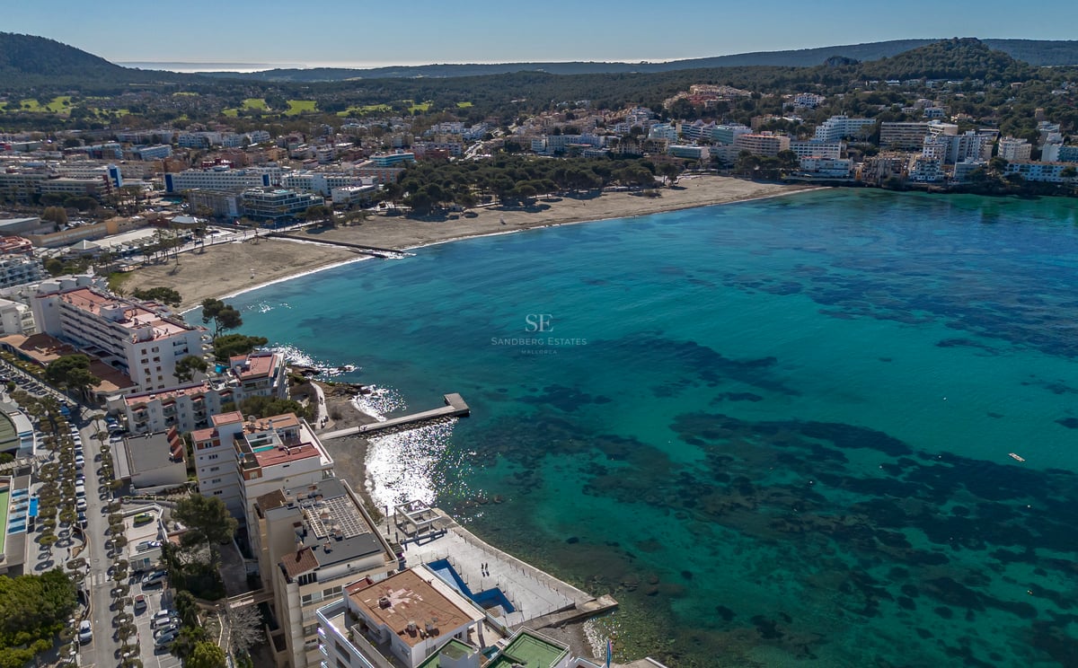 Vue aérienne par drone d'une baie turquoise avec plage de sable et bâtiments côtiers sous un ciel bleu clair.