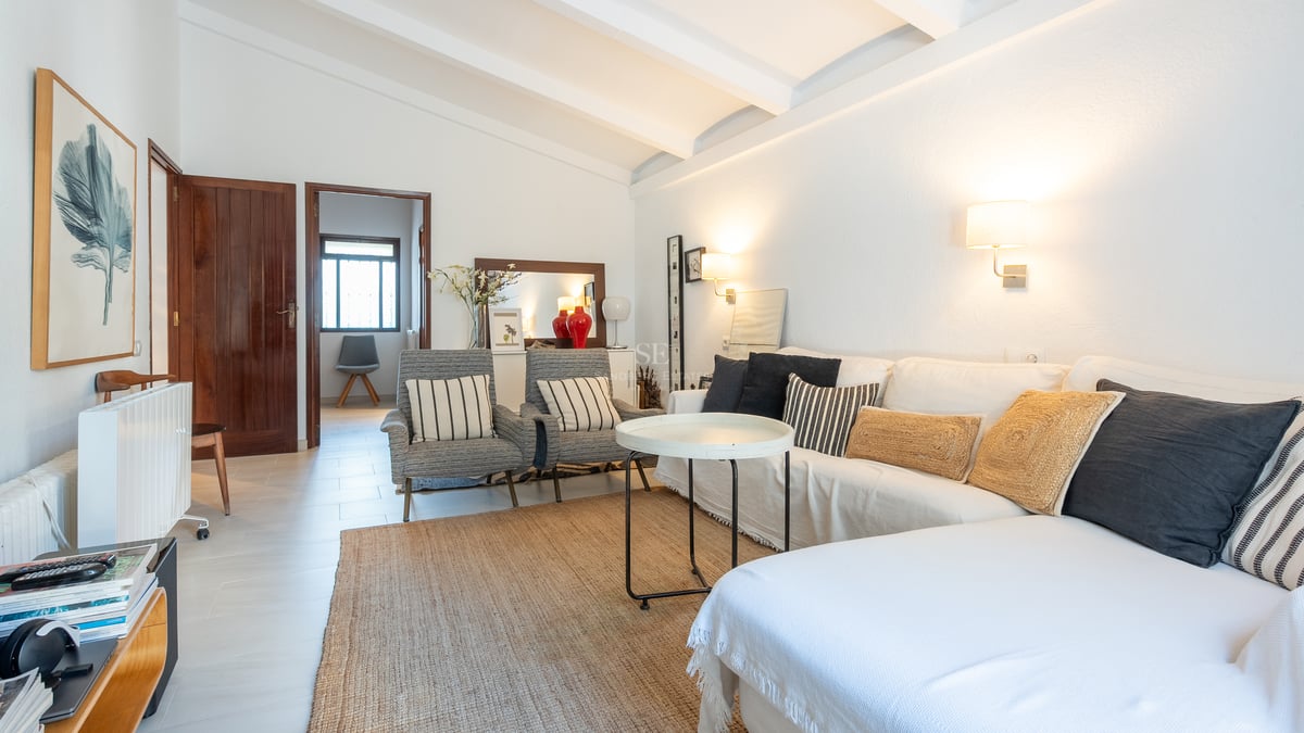 Salon lumineux avec canapé blanc, fauteuils rayés, tapis en jute et poutres de plafond blanches.