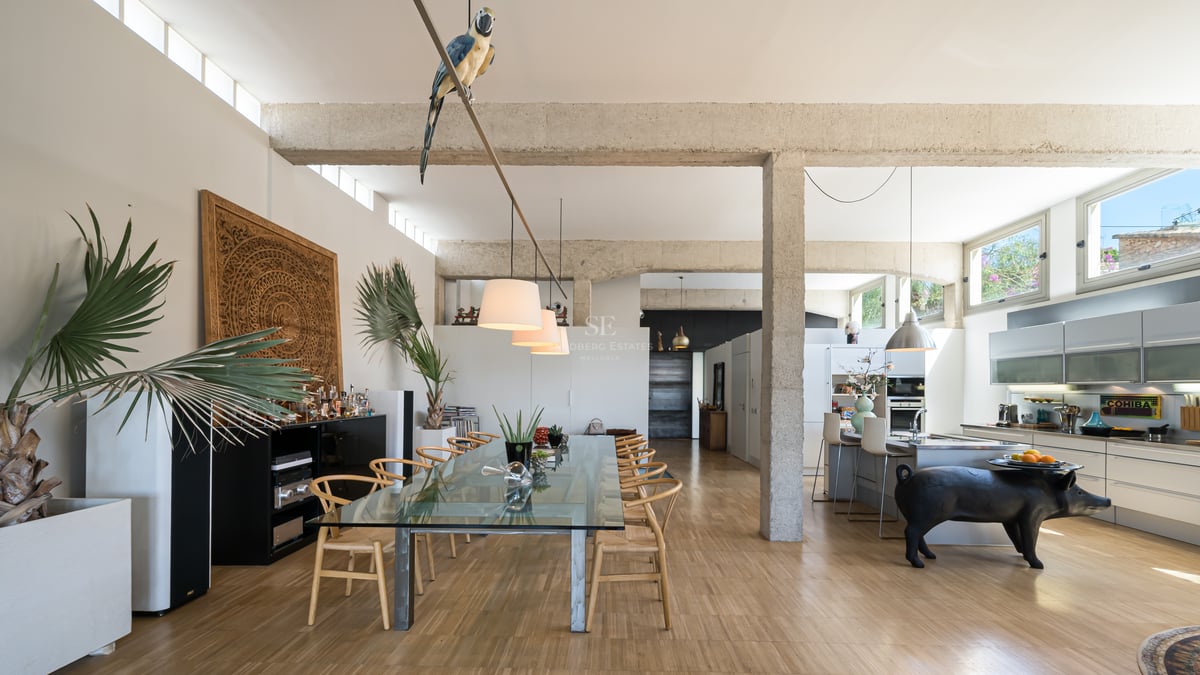 Vaste salle à manger avec poutres en béton brut, table en verre et cuisine intégrée baignée de lumière.
