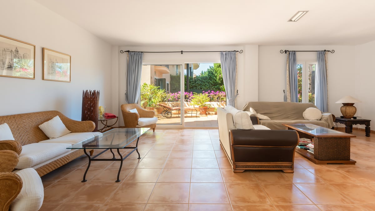 Grand salon avec canapés en osier, sol en terre cuite et grandes baies vitrées ouvrant sur une terrasse ensoleillée.
