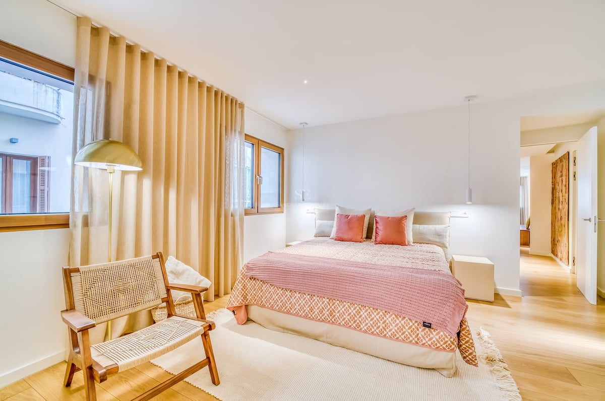Chambre lumineuse avec lit double, textiles roses, parquet en bois, fauteuil en osier et fenêtres avec rideaux.