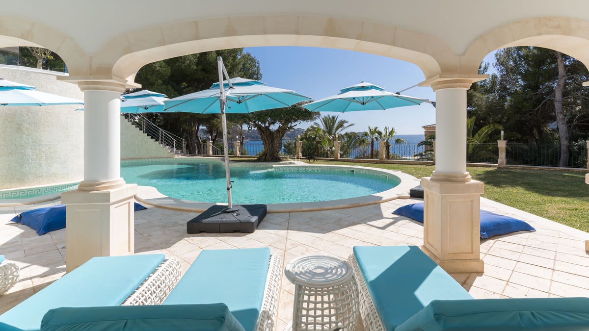 Une piscine turquoise incurvée entourée de dalles en pierre et de chaises longues turquoises sous une arche ombragée.
