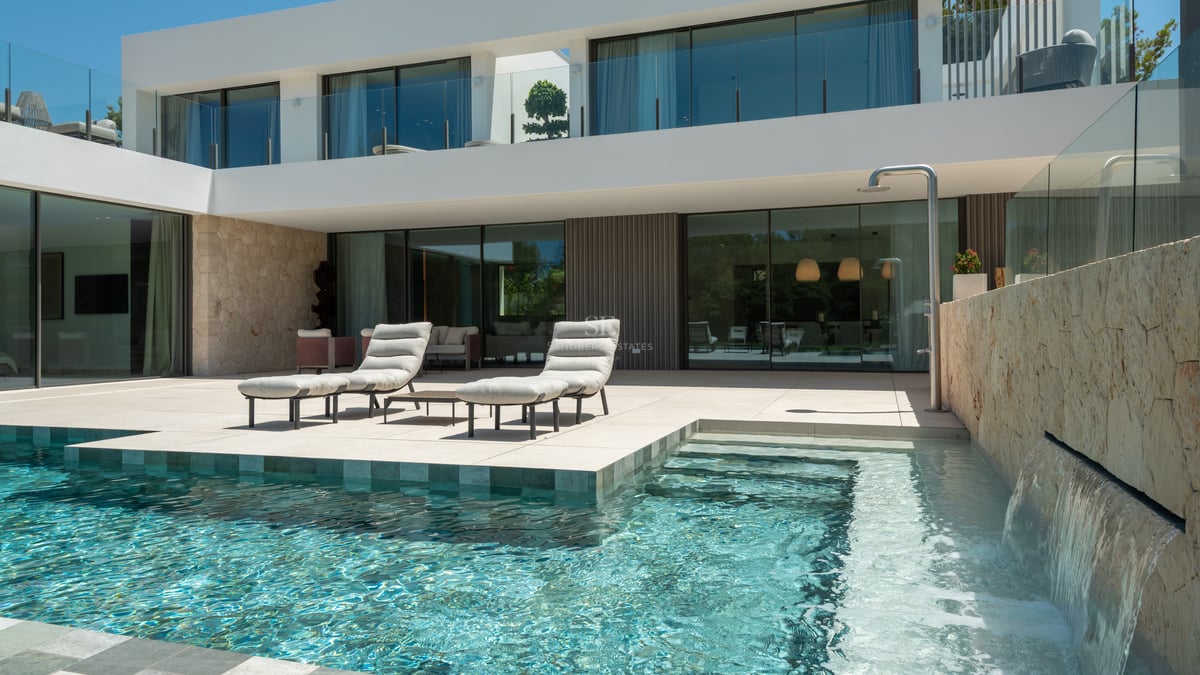Terrasse de villa moderne avec piscine turquoise, cascade en pierre, bains de soleil et baies vitrées.