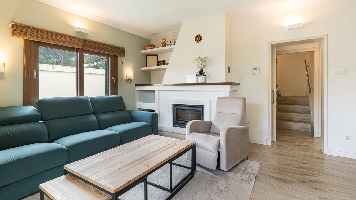 Salon moderne avec canapé bleu canard, cheminée blanche, table basse en bois et fauteuil beige.