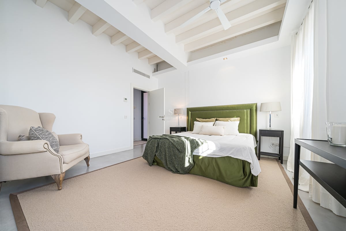 Chambre lumineuse avec poutres blanches, lit en velours vert, fauteuil beige et grandes fenêtres.