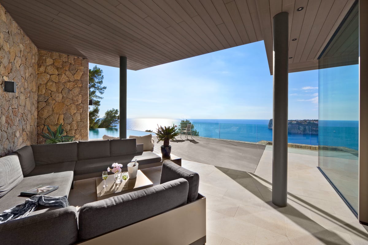 Salon extérieur avec canapés gris sur une terrasse en pierre surplombant une piscine à débordement et la mer.