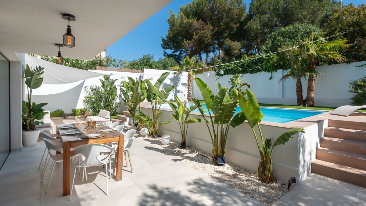 Une terrasse moderne avec table à manger en bois sous un auvent à côté d'une piscine surélevée et de plantes tropicales.