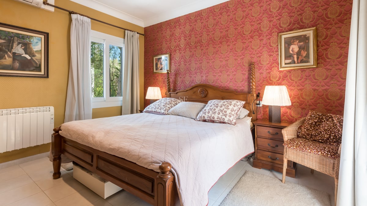 Chambre traditionnelle avec lit en bois, papier peint damas rouge et fenêtre donnant sur des arbres.