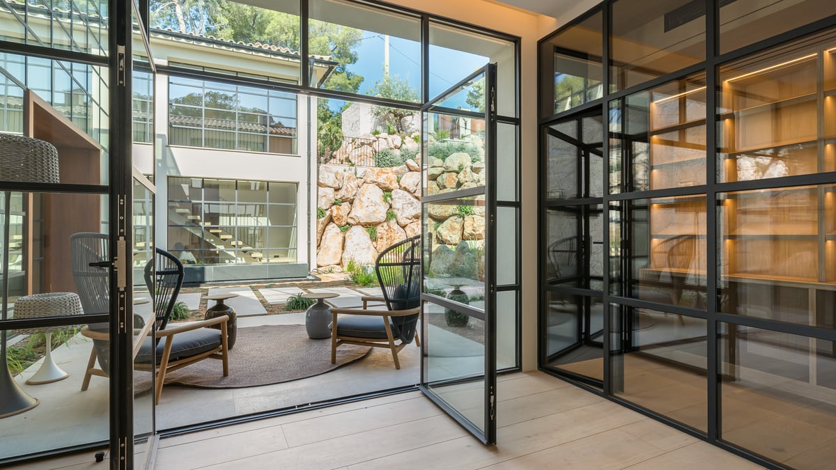 Vue intérieure vers une terrasse avec portes en acier noir, fauteuils design et mur en pierre naturelle.