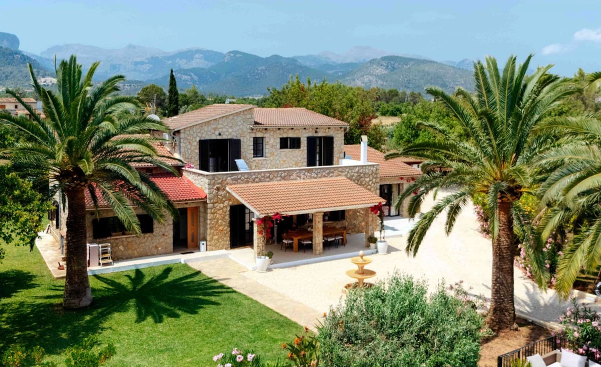 Villa traditionnelle en pierre avec palmiers, pelouse verdoyante et montagnes en arrière-plan.