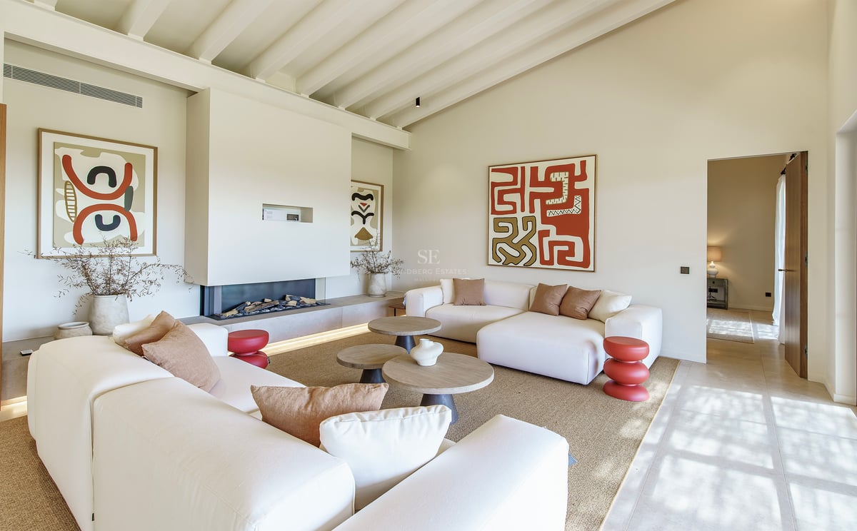 Salón luminoso con techos altos de vigas, chimenea moderna integrada, sofás blancos y arte abstracto en tonos tierra.