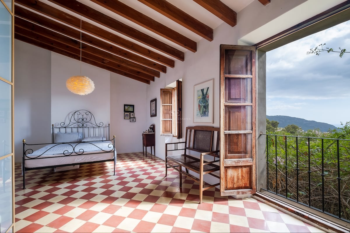 Chambre avec sol en damier, lit en fer forgé, poutres en bois et porte ouverte sur des montagnes verdoyantes.