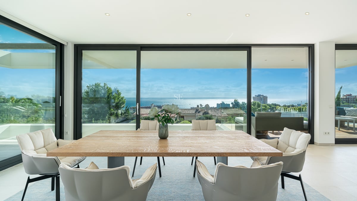 Salle à manger lumineuse avec table en bois et chaises grises face à des baies vitrées avec vue sur l'océan.