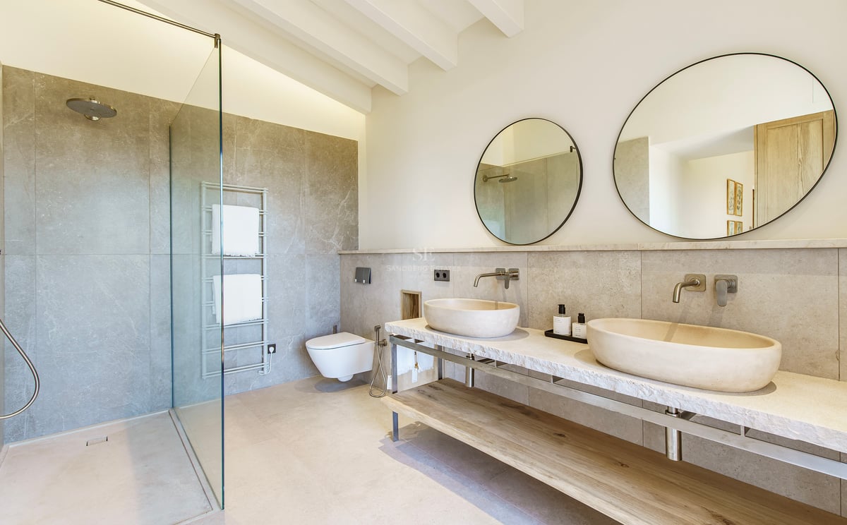 Baño de lujo con dos lavabos de piedra sobre encimera de mármol, espejos circulares y ducha de cristal.