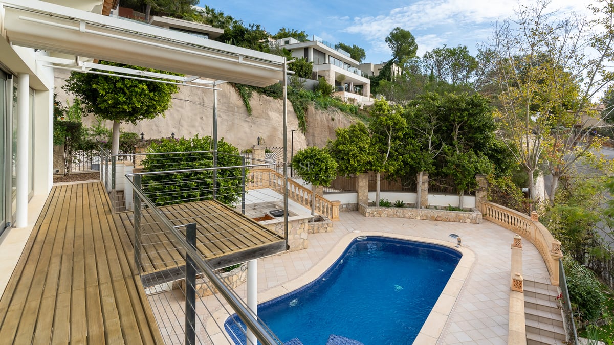 Vue plongeante sur une piscine bleue, une terrasse en bois et un patio carrelé entourés de verdure.
