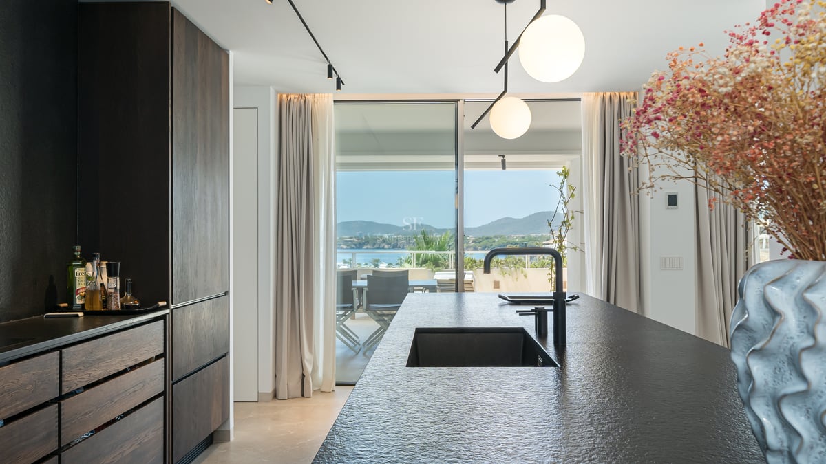 Cuisine moderne avec un îlot en pierre sombre et un évier noir donnant sur une terrasse ensoleillée et la mer.