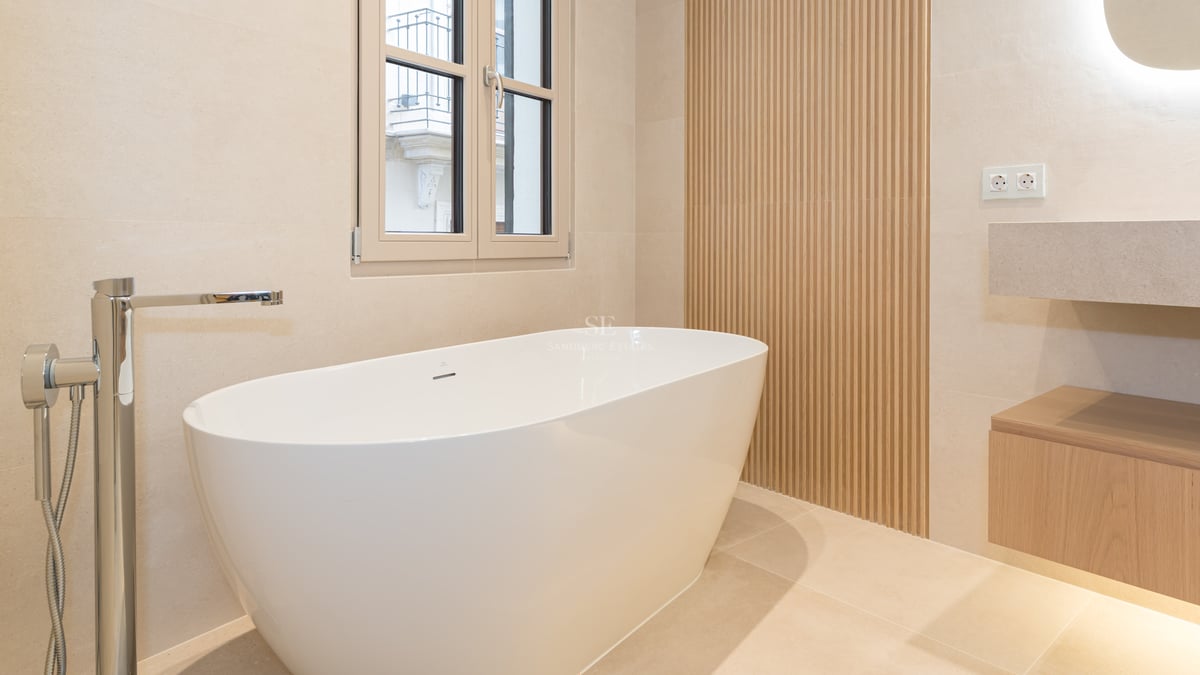 Modern white freestanding bathtub next to a light wood slatted wall and chrome floor faucet on beige tiles.