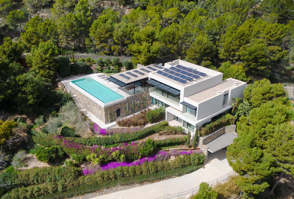 Vue aérienne d'une villa moderne blanche avec piscine à débordement, panneaux solaires et forêt de pins.