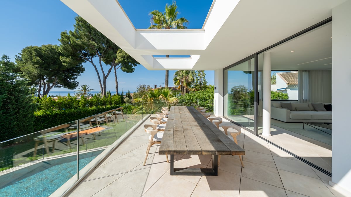 Terrasse blanche moderne avec grande table en bois, garde-corps en verre et vue sur la mer et la piscine.