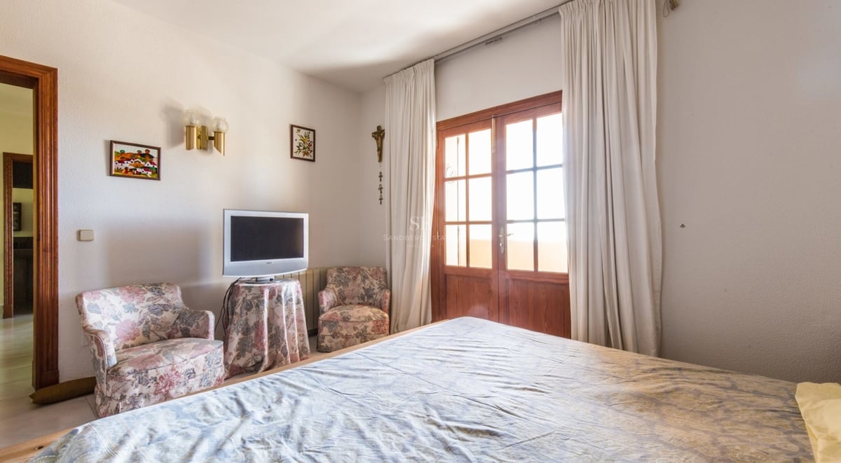 Chambre avec lit, deux fauteuils à fleurs et portes en bois ouvrant sur un balcon.