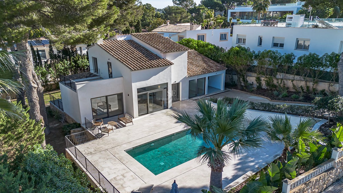 Vue aérienne d'une villa blanche avec toit en tuiles, piscine turquoise et grande terrasse entourée de pins et palmiers.