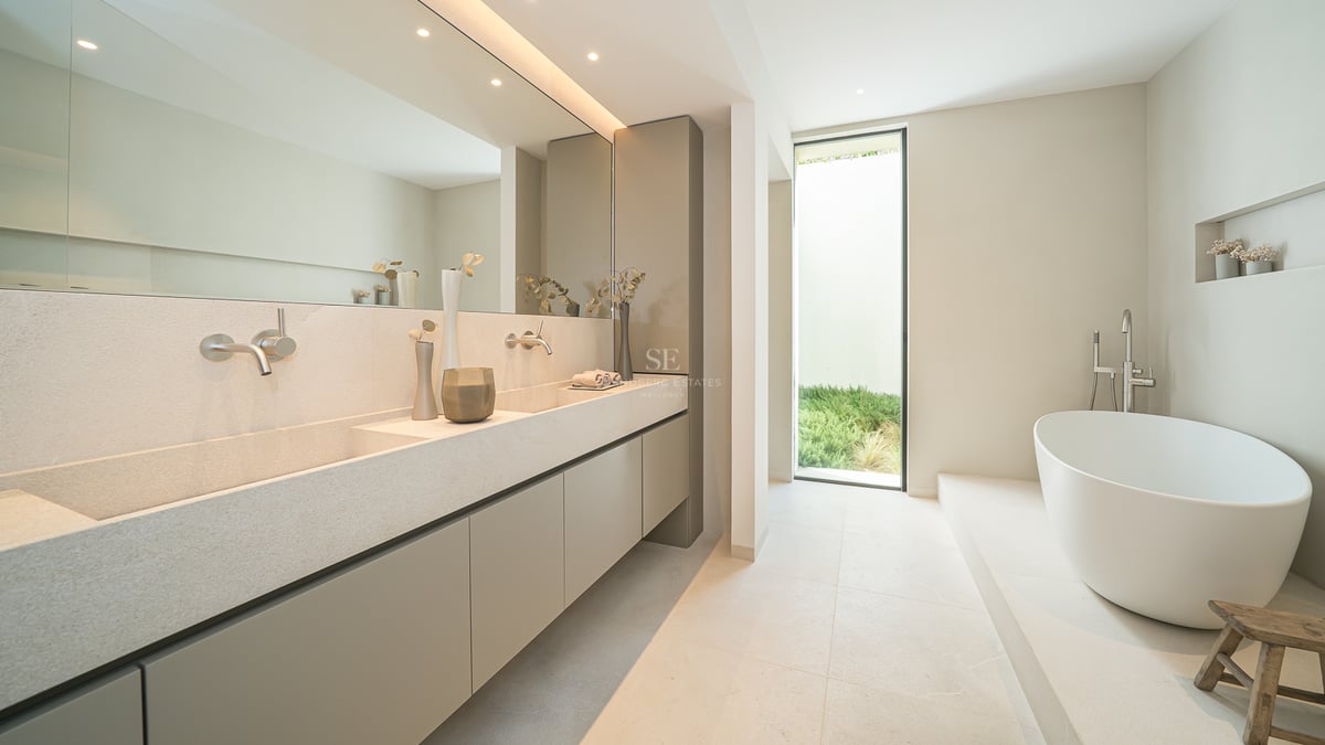 Salle de bain minimaliste avec double vasque en pierre, grand miroir, baignoire blanche et fenêtre sur le jardin.