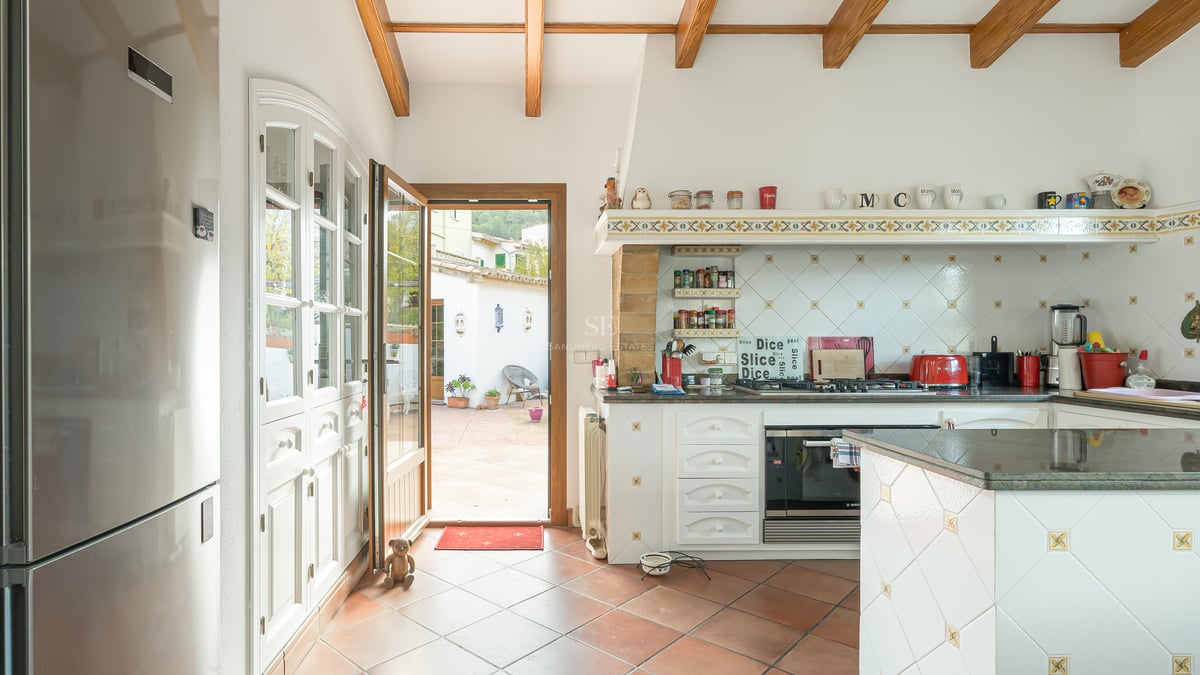 Cuisine avec poutres en bois, sol en terre cuite et porte ouverte sur une cour ensoleillée.