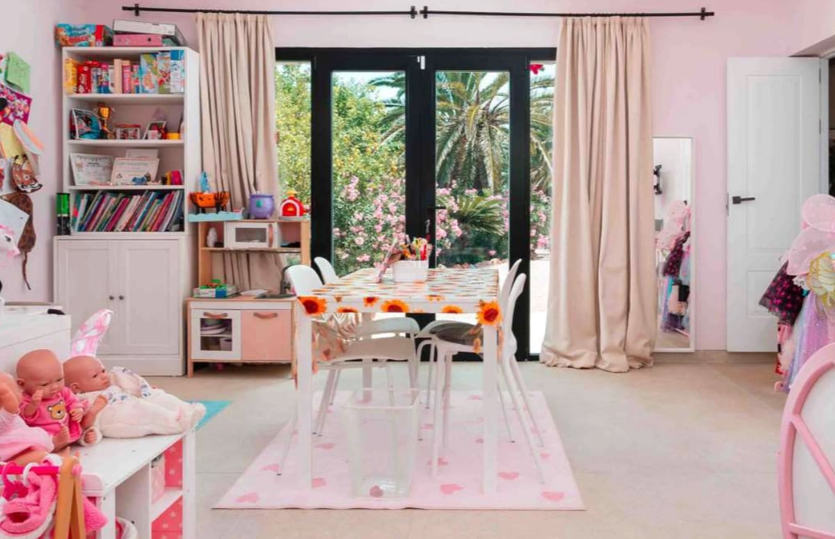 Une salle de jeux pour enfants aux murs roses, dotée de baies vitrées ouvrant sur un jardin avec palmiers.