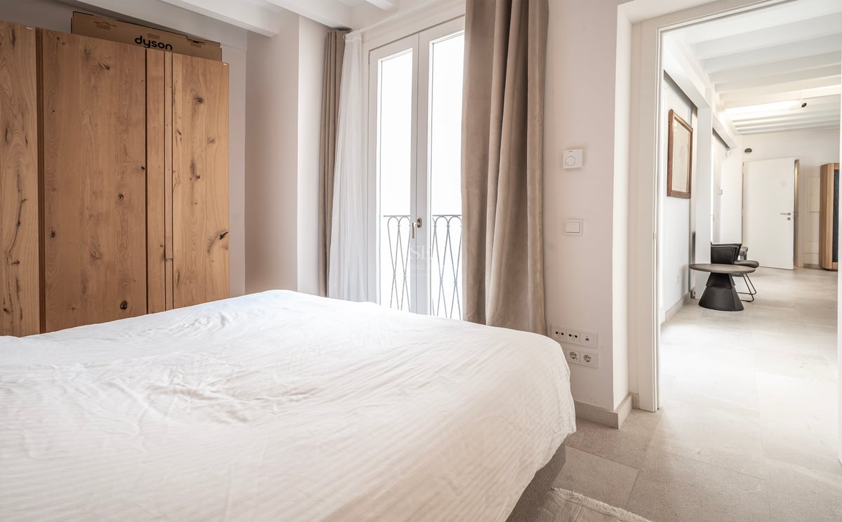 Chambre lumineuse avec lit blanc, grande armoire en bois et portes-fenêtres donnant sur un balcon.