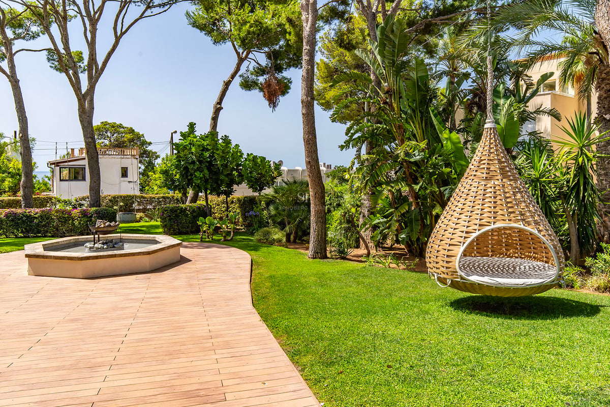Jardin luxuriant avec terrasse en bois, pins parasols et fauteuil suspendu design sur une pelouse entretenue.