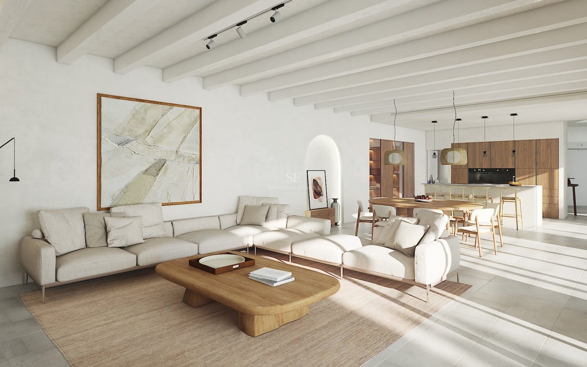 Salón luminoso de planta abierta con vigas blancas, sofá modular, mesa de madera y cocina integrada al fondo.