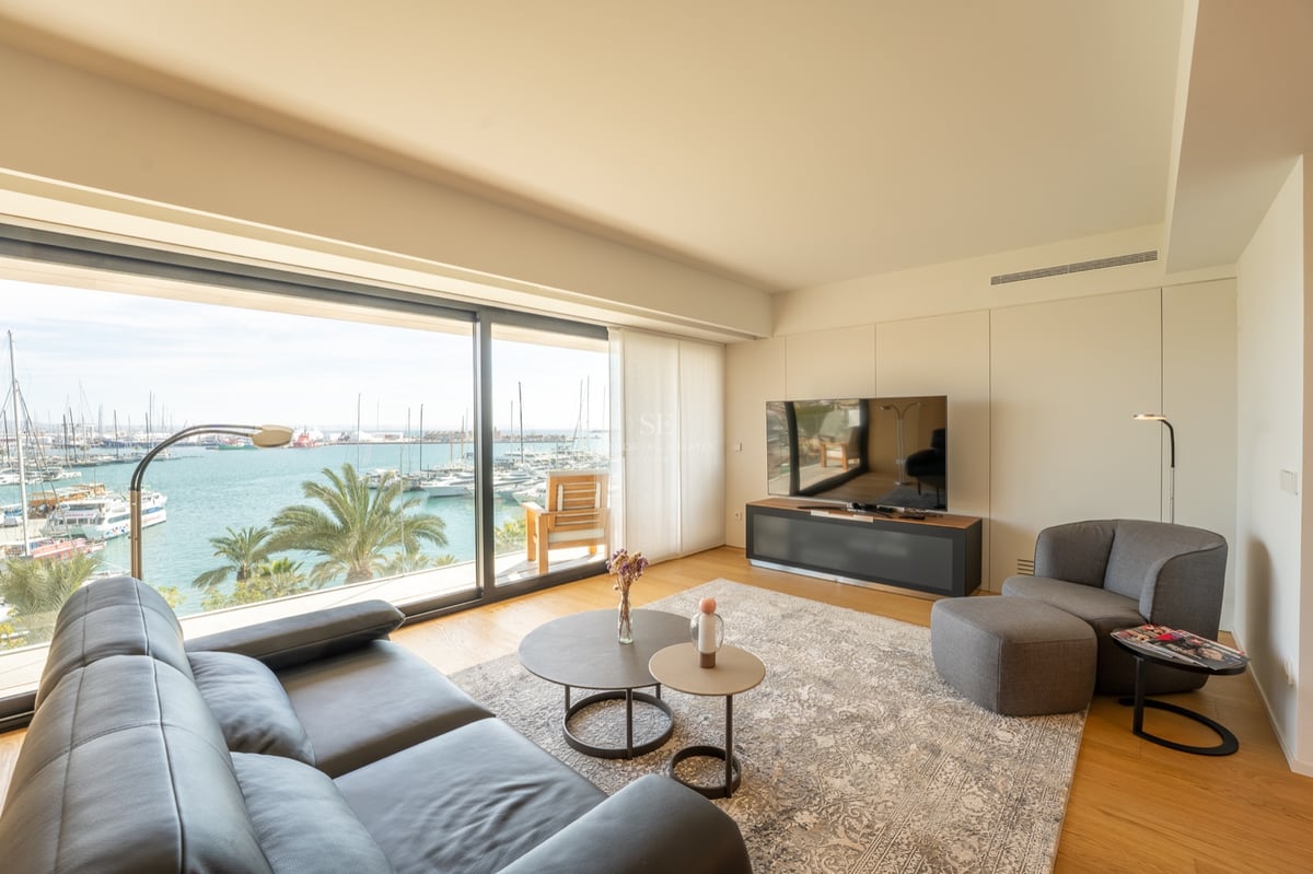 Salon moderne avec canapé en cuir noir et grandes baies vitrées donnant sur une marina ensoleillée.