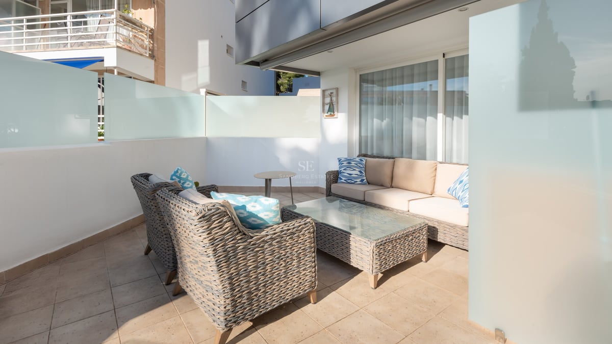 Terrasse moderne avec canapés en osier gris, coussins beige et bleus sur un patio ensoleillé avec parois en verre dépoli.