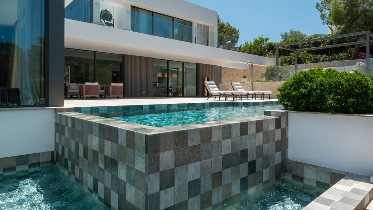 Villa blanche moderne avec piscine à débordement en pierre, chaises longues et grandes baies vitrées.
