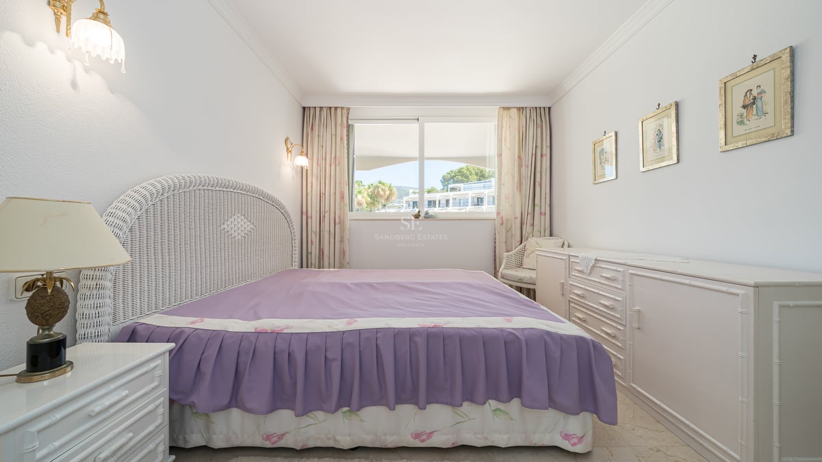 Chambre avec tête de lit en osier blanc, couvre-lit violet et fenêtre offrant une vue sur l'extérieur.
