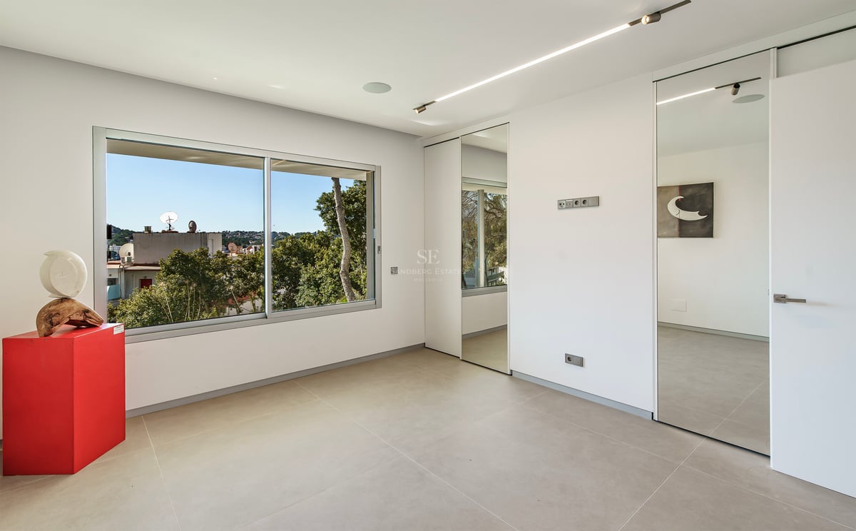 Chambre moderne avec sol gris, placards en miroir et grande fenêtre donnant sur la verdure.
