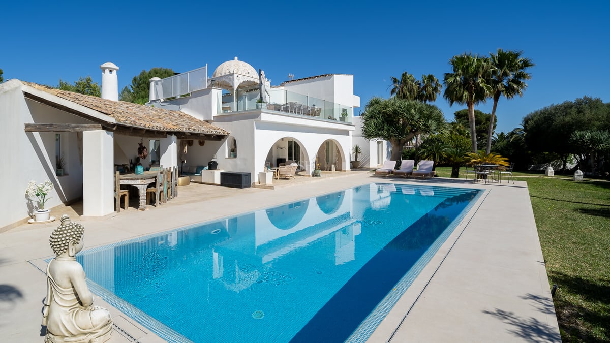Une grande piscine bleue reflétant une villa méditerranéenne blanche avec des arches, des palmiers et une statue de Bouddha.