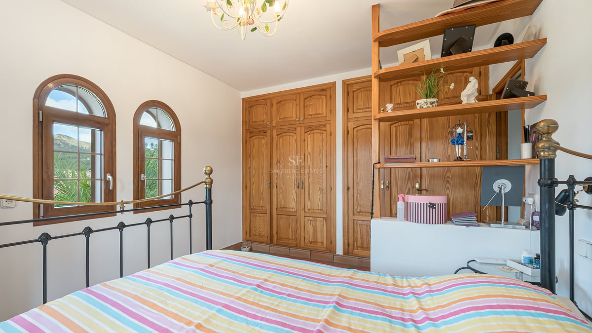 Chambre avec lit en fer forgé, armoires en bois intégrées et fenêtres cintrées offrant une vue sur le paysage.