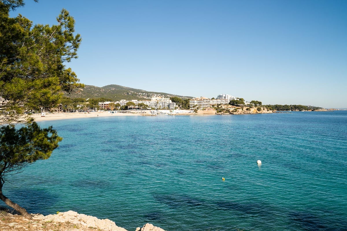 Vue panoramique d'une baie méditerranéenne aux eaux turquoises, plage de sable et bâtiments blancs sous un ciel bleu.