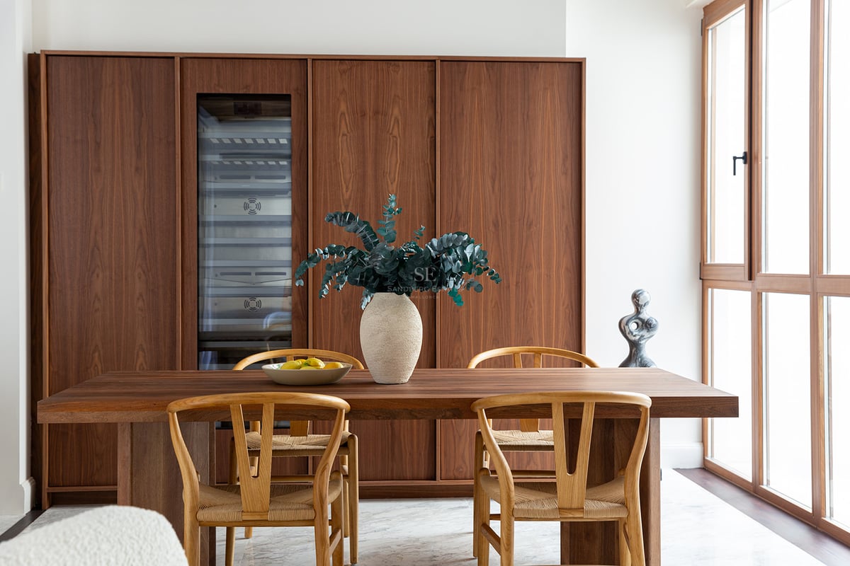 Comedor moderno con armarios de nogal, vinoteca, mesa de madera, sillas wishbone y suelos de mármol blanco.
