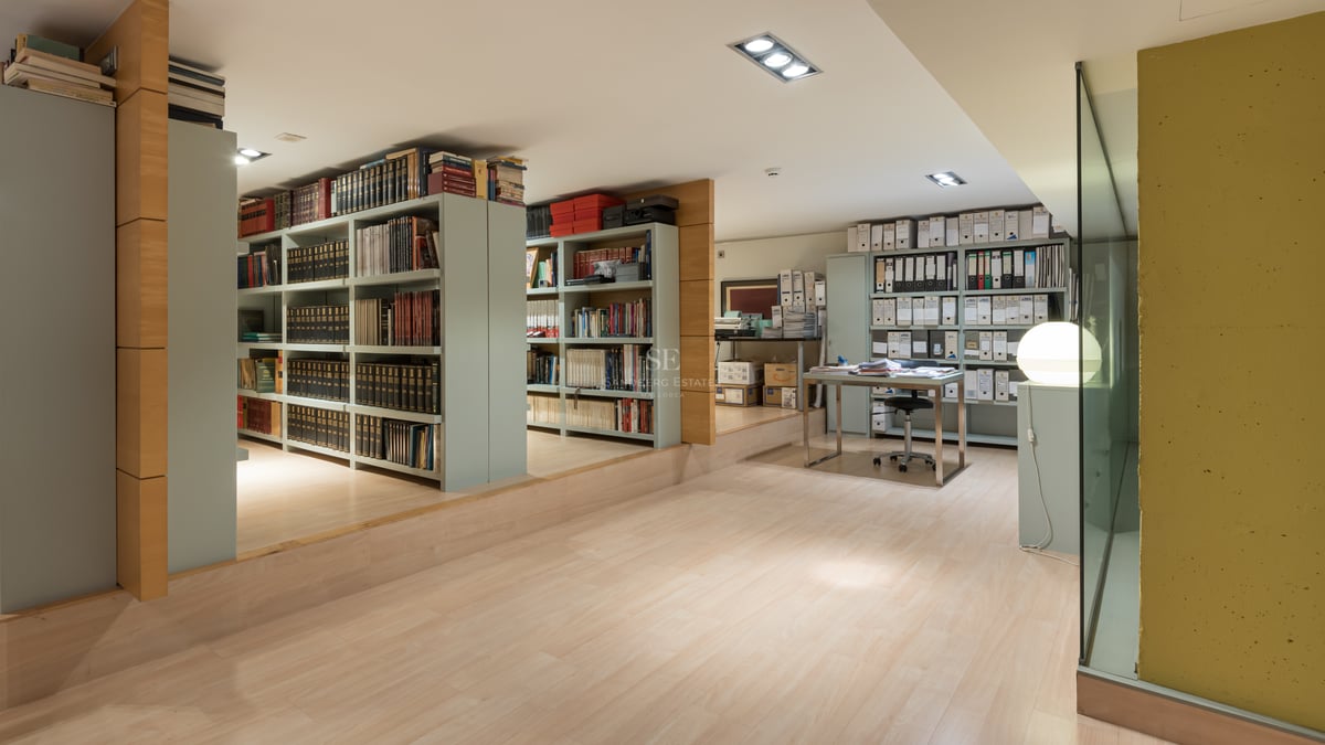 Spacious home office with extensive book shelving, light wood floors, and a modern glass-top desk.