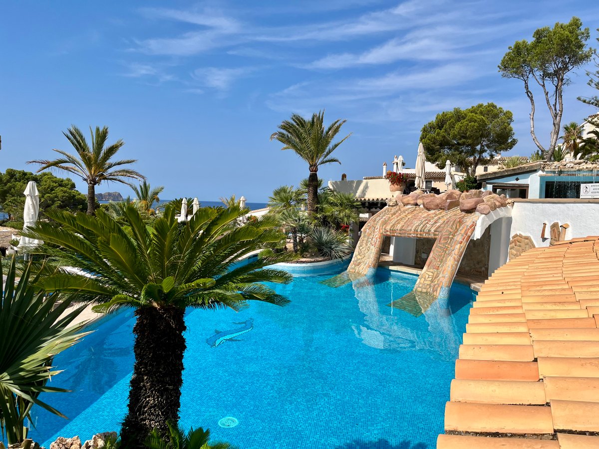 Grande piscine bleue entourée de palmiers et d'une architecture méditerranéenne avec l'océan à l'horizon.