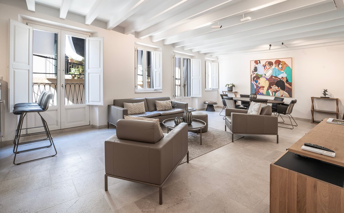 Salon lumineux avec canapés en cuir gris, plafond aux poutres blanches et coin repas avec une œuvre d'art colorée.