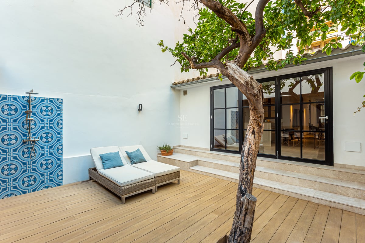 Terrasse en bois avec transats, douche extérieure aux carreaux bleus et un arbre devant de grandes baies vitrées.