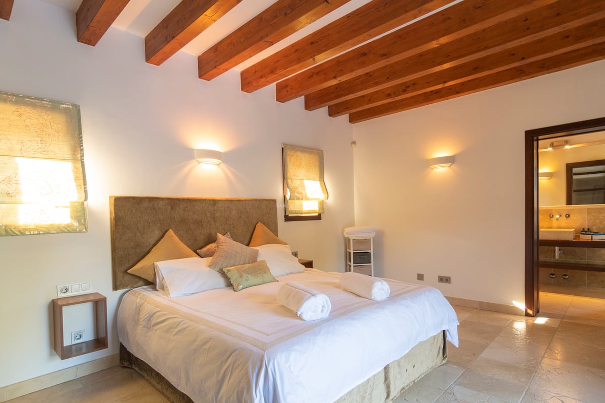 Chambre à coucher avec poutres en bois sombre, sol en pierre et salle de bain attenante.