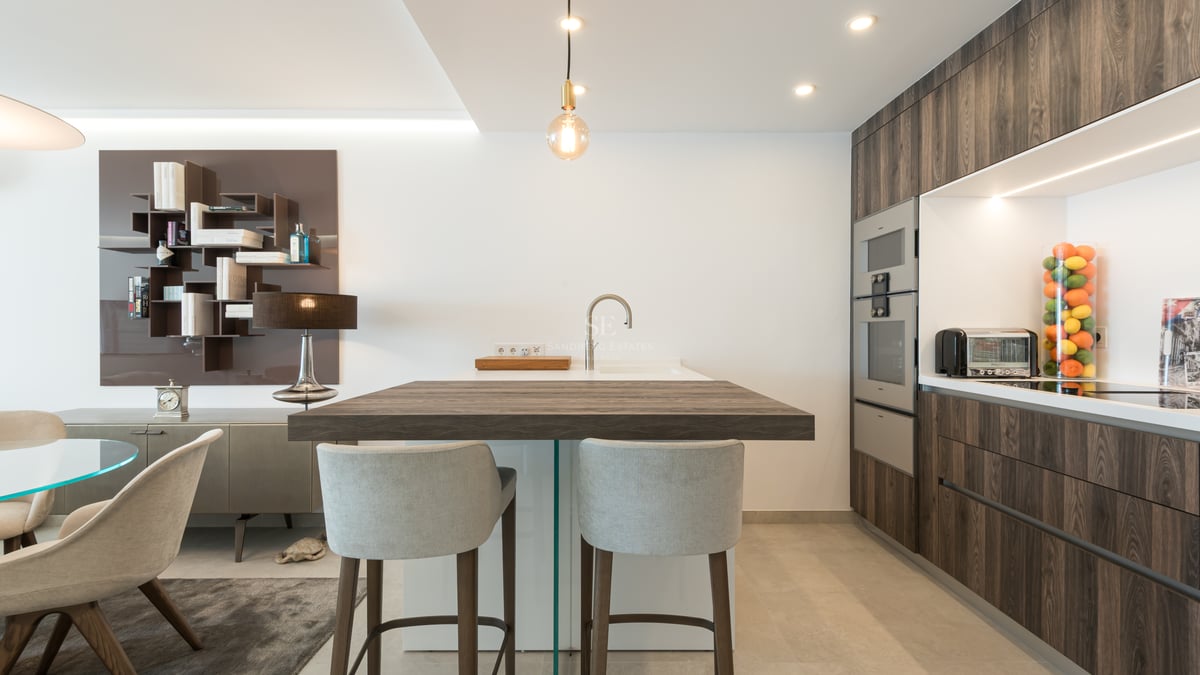 Cuisine moderne avec îlot en bois foncé, tabourets gris, armoires épurées et appareils électroménagers intégrés.