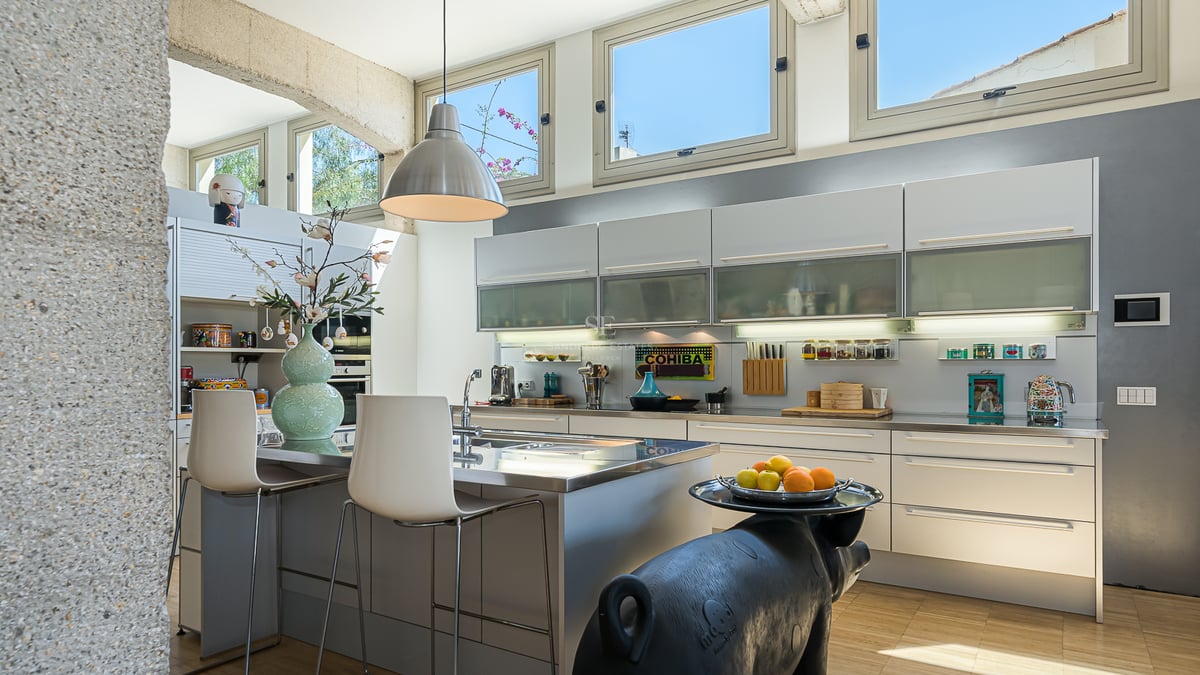 Cuisine moderne avec îlot en acier inoxydable, placards blancs et fenêtres hautes sous une arche en pierre.